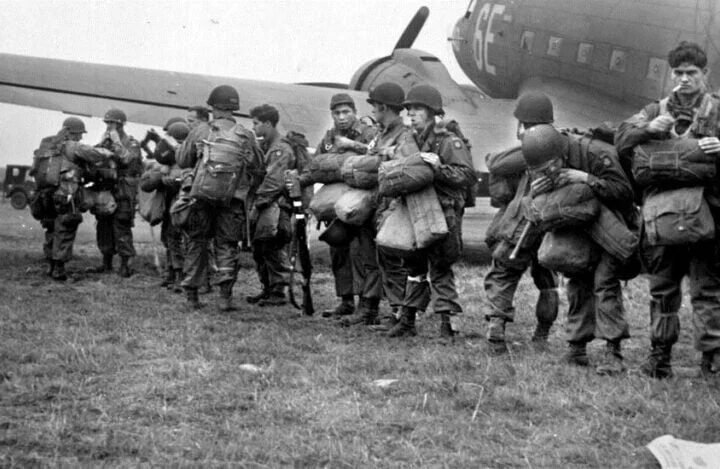 Tropas paraquedistas aliadas preparando-se para a Operação Market Garden em 16 de setembro de 1944, com aeronaves de transporte e equipamentos militares ao fundo.