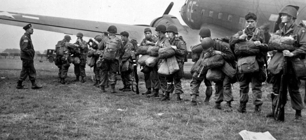 Tropas paraquedistas aliadas preparando-se para a Operação Market Garden em 16 de setembro de 1944, com aeronaves de transporte e equipamentos militares ao fundo.